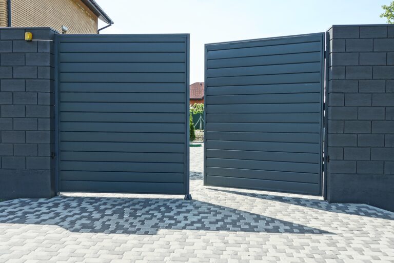 one open black metal gate and part of a brick wall of a fence on the street on a gray sidewalk