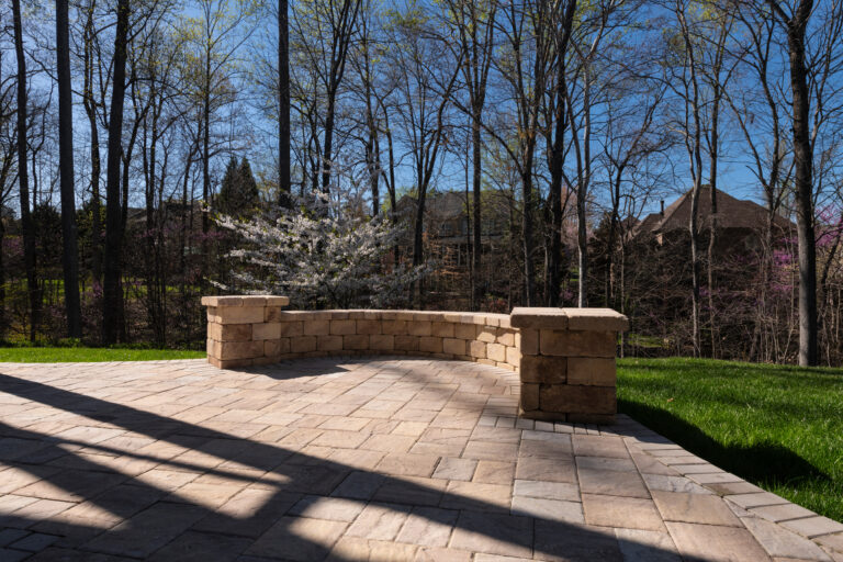 Picturesque backyard view in spring season with patio pavers and stone wall, blooming white cherry tree, and spring colored woods in the background.